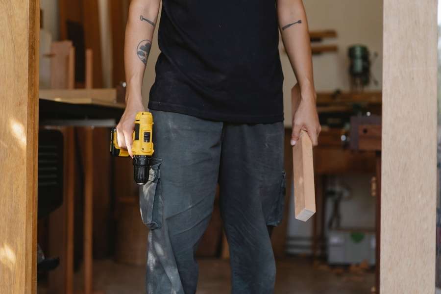 A carpenter holding a power drill and a wood piece in a workshop, ready to work and avoid common joinery mistakes.