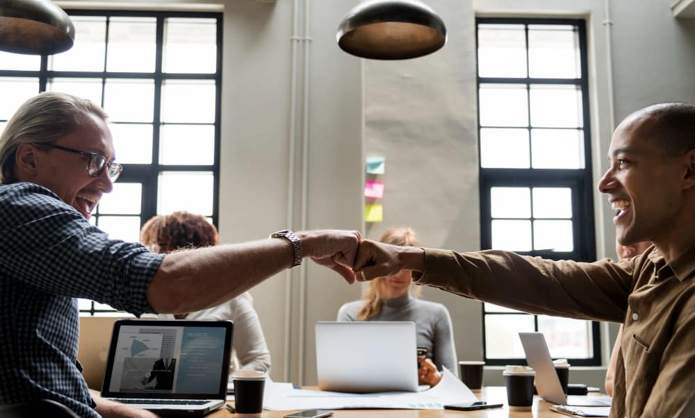 Two smiling colleagues fist-bumping across a meeting table in a bright, modern office, demonstrating teamwork and encouraging creativity and collaboration.