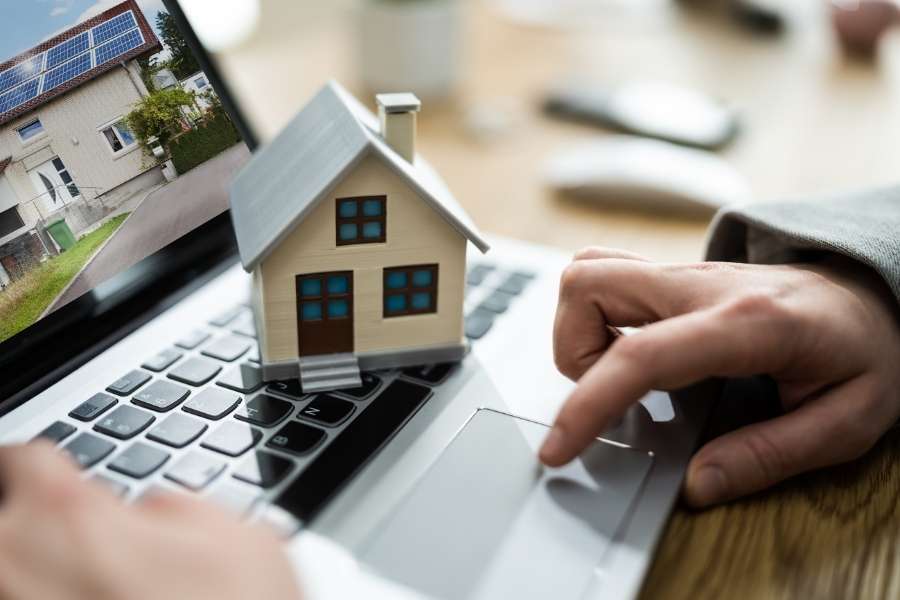 A person using a laptop to browse a property listing with a miniature house model placed on the keyboard.