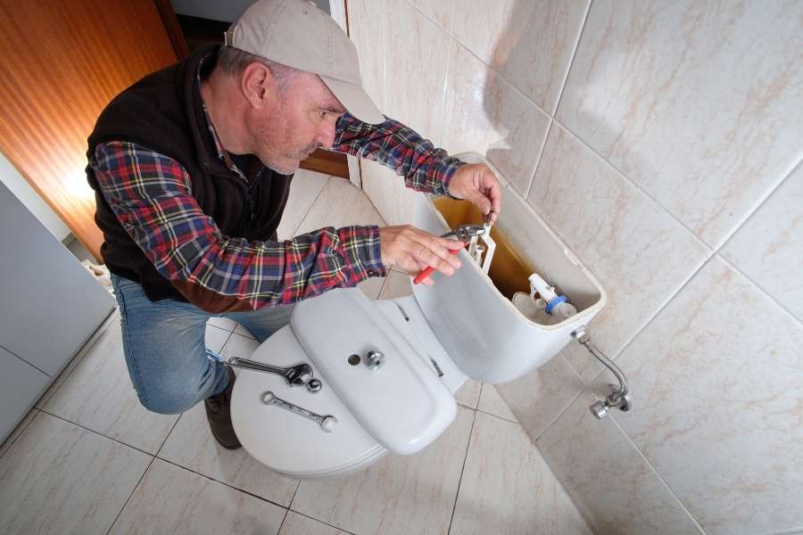 A plumber kneeling down to fix a running toilet by adjusting mechanisms inside the tank.