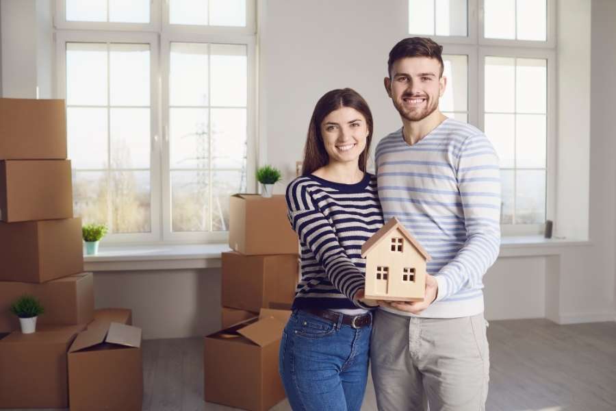 A happy couple holding a model house in an empty room with moving boxes to secure a mortgage.
