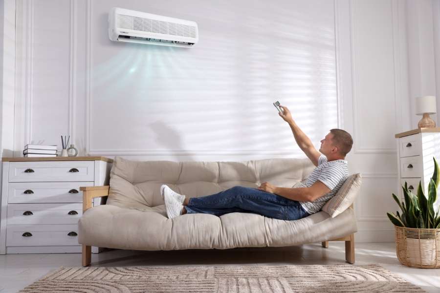 A man relaxing on a beige sofa in a modern living room while using a remote to control an energy-efficient air conditioning unit mounted on the wall.